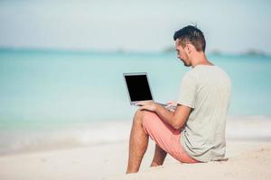 joven sentado en la arena con una computadora portátil en una playa caribeña tropical. hombre con computadora y trabajando en la playa foto