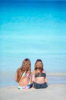 Adorable little girls playing with sand on the beach. Back view of kids sitting in shallow water and making a sandcastle photo