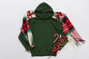 Close up green blank template hoodie copy space. Christmas Holiday concept. Top view mockup hoodie, scarf, hat. Red holidays decorations white background. Happy New Year accessories. Selective focus photo