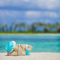 Straw bag, blue hat, sunglasses and sunscreen bottle on tropical beach photo