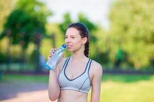 Young woman drinking water after running outside. Female fitness model training outside in the park. Concept of healthy and proper nutrition. photo