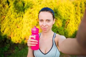 mujer joven con botella de agua después de correr afuera. modelo de fitness femenino entrenando afuera y tomando selfie en el parque. estilo de vida de fitness de bienestar saludable. foto