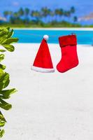 sombrero rojo de santa y calcetín de navidad colgando en la playa tropical. concepto de navidad. foto