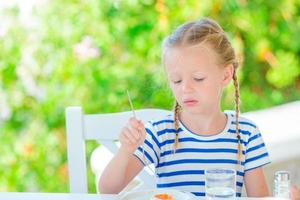 Adorable little girl having breakfast at cafe with sea view early in the morning photo