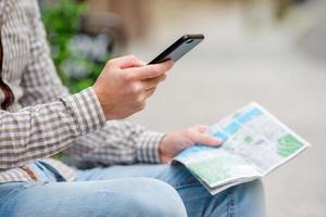 Closeup of male hands holding cellphone and city map outdoors on the street. Man using mobile smartphone to find famous attraction. photo