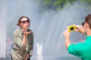 Young man taking a picture of his girlfriend while sitting background the fountain. Young man making photo of woman on the street laughing and having fun in summer.