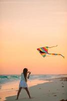 Little running girl with flying kite on tropical beach. Kid play on ocean shore. photo