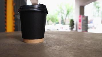 une tasse de papier noir avec du café sans logo sur une table de café de rue avec des gens méconnaissables marchant sur un fond flou tôt le matin par une journée ensoleillée. video