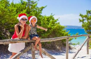 madre joven y niña con sombrero rojo de navidad en la playa tropical foto