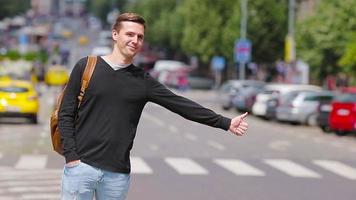 junger glücklicher mann nimmt ein taxi in europäischen straßen. porträt eines kaukasischen touristen mit rucksack, der lächelt und ein taxi nimmt video