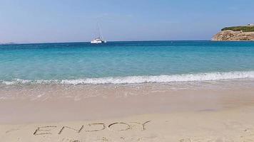 Word Enjoy handwritten on sandy beach with soft ocean wave on background. Idea for advertising video