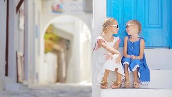 twee meisjes in blauw jurken hebben pret buitenshuis. kinderen Bij straat van typisch Grieks traditioneel dorp met wit muren en kleurrijk deuren Aan Mykonos eiland, in Griekenland video
