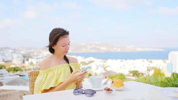 Schönes Mädchen beim Frühstück mit Telefon im Café im Freien mit herrlichem Blick auf die Stadt Mykonos. video