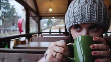 una bella donna in abiti casual, un maglione grigio e un cappello lavorato a maglia si siede in un bar vicino alla finestra e beve il caffè mattutino. tazza verde con bevanda calda in mani femminili video