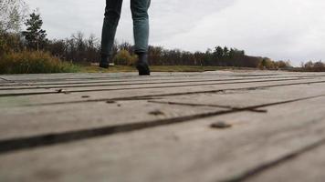 una mujer caminando con botas, jeans y una chaqueta en un puente de madera sobre un lago en otoño, un alegre viaje feliz sin preocupaciones. relajación, disfrutar de la vida en la naturaleza, ángulo de visión bajo. video