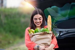 Caucasian brunette going holding paper bags with food products. Young woman putting package with groceries and vegetables into car trunk. Attractive caucasian female shopping in mall or grocery store photo