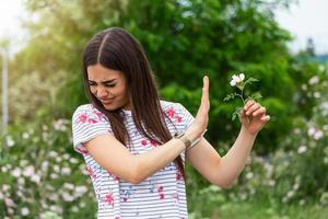 Young woman with Pollen allergy holding a flower and saying no.. Young woman with pollen and grass allergies. Flowering trees in background. Spring Seasonal allergies and health problems. photo