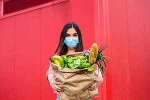 Young woman in medical mask holds an eco paper bag with food, fruits and vegetables, pepper, baguette, lettuce, safe online smart delivery, coronovirus, quarantine, pandemic, stay home concept photo