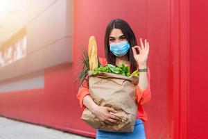 Woman with medical mask holding a shopping bag full of fresh food. Young woman with a grocery shopping bag during covid 19, coronavirus pandemic . Beautiful young woman with vegetables in grocery bag. photo