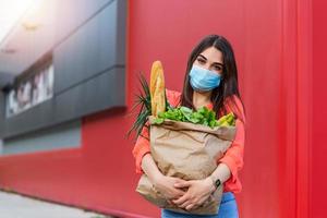 Buyer wearing a protective mask.Shopping during the Covid 19, Coronavirus pandemic quarantine. Woman in medical mask holds a paper bag with food, fruits and vegetables photo