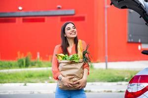 mujer joven montando carrito de compras lleno de comida en el estacionamiento al aire libre. mujer joven en el aparcamiento, cargando las compras en el maletero del coche. Compra realizada con éxito. mujer poniendo bolsas en el coche después de ir de compras foto