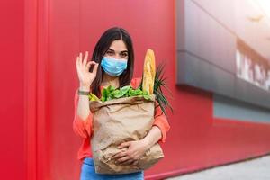 Woman with medical mask holding a shopping bag full of fresh food. Young woman with a grocery shopping bag during covid 19, coronavirus pandemic . Beautiful young woman with vegetables in grocery bag. photo