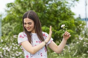 Young woman with Pollen allergy holding a flower and saying no.. Young woman with pollen and grass allergies. Flowering trees in background. Spring Seasonal allergies and health problems. photo