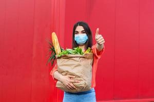 Pretty girl with medical mask holding bag with groceries and looking at camera with thumb up. Woman holding heavy bag with groceries. Groceries shopping during Covid 19, coronavirus pandemic