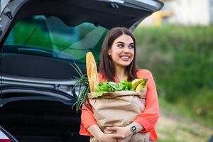 hermosa joven comprando en un supermercado, poniendo los comestibles en su auto en el estacionamiento. mujer después de ir de compras y conducir a casa ahora con su coche al aire libre. foto