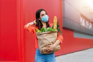 Woman with medical mask holding a shopping bag full of fresh food. Young woman with a grocery shopping bag during covid 19, coronavirus pandemic . Beautiful young woman with vegetables in grocery bag. photo