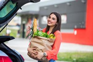 Caucasian brunette going holding paper bags with food products. Young woman putting package with groceries and vegetables into car trunk. Attractive caucasian female shopping in mall or grocery store photo