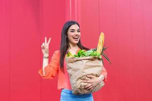 Beautiful young woman smiling holding a paper bag full of groceries. Happy pretty girl holding bag with groceries over red background photo