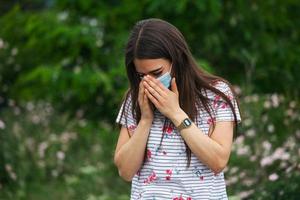 Portrait of young woman wearing a protective mask sneezes on green nature background with flowers in protective medical mask, coronavirus pandemic photo