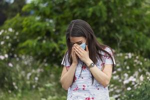 Portrait of beautiful sad woman in medical mask sneezes among white flowers. coronavirus pandemic. Concept of not spread of the virus. photo