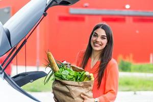 Woman after shopping in a mall or shopping centre and driving home now with her car outdoor. Beautiful young woman shopping in a grocery store supermarket, putting the groceries into her car photo