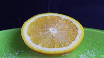 una rodaja redonda de naranja que se ve muy jugosa y apetitosamente espolvoreada con azúcar. tomas de primer plano de azúcar en una rodaja de cítricos recién cortada en un plato verde, sobre un fondo negro. video