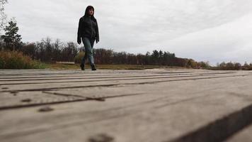 una mujer caminando con botas, jeans y una chaqueta en un puente de madera sobre un lago en otoño, un alegre viaje feliz sin preocupaciones. relajación, disfrutar de la vida en la naturaleza, ángulo de visión bajo. video