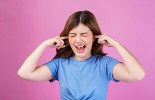 Portrait of angry irritated young woman wearing casual t-shirt covering her ears with hands and shouting while standing isolated over pink background photo