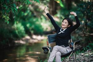 mujer joven feliz sentada en una silla mientras acampa en el bosque foto