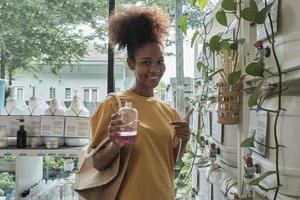 A Black female customer fills liquid soap into recycling jar from reusable containers in a zero-waste and refill store, smiles, and looks at camera, happy shopping in an environment-friendly shop. photo
