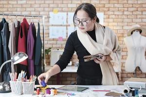 Asian middle-aged female fashion designer works in studio by choosing thread and fabric color collection ideas and style concepts for dress design jobs. Professional boutique tailor SME entrepreneur. photo