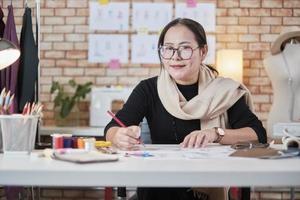 Asian middle-aged female fashion designer working in a studio by idea drawing sketches with colorful thread and sewing for a dress design collection, professional boutique tailor SME entrepreneur. photo