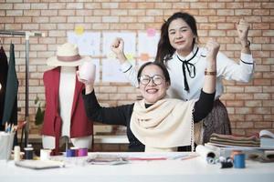 Asian middle-aged female fashion designer and young teen trainee tailor celebrate and cheerful with dress design work success and satisfy in stylist studio, happy costume boutique small business. photo