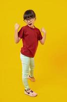Portrait of Asian pretty girl celebrating and raising hand up for successful, Thai kid with glasses in red shirt have glad and joy on yellow in studio photo