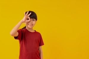 Young Asian girl kid in red shirt making ok sign with hand on eye looking through fingers on yellow background in studio photo