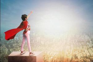 super niña muestra poder en la azotea del edificio, niña pequeña con capa de héroe rojo muestra la mano hacia el cielo y la ciudad en puesta de sol, sueño activo y concepto de idea de motivación