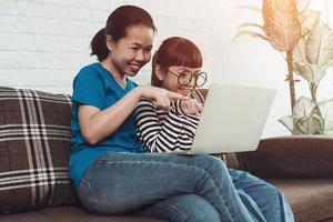 Young Asian girl kid with mom use laptop on sofa. Learning and searching internet, family activity at home photo