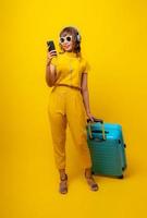 Cute and funny woman in yellow casual cloth style holding cell phone, wearing ear headphones and dragging blue luggage or suitcase in studio background