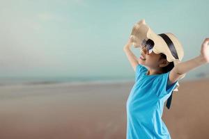 Young pretty Asian kid with travel bag in summer style, happy Thai girl wearing hat and sunglasses looking sea and beach in vacation