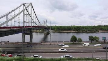 vista del puente peatonal para bicicletas que cruza el río dnieper hasta la isla trukhanov y la carretera con coches en movimiento a lo largo del paseo marítimo. nublado. centro de la ciudad. ucrania, kiev - 14 de junio de 2020. video